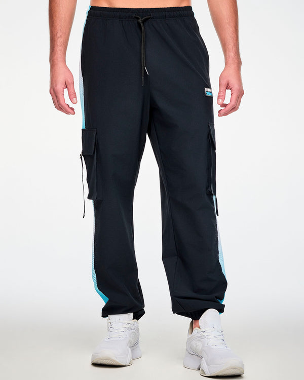 Male model in black woven cargo pants with light blue ombre mesh side panels, a drawcord waist, elastic cuffs, and a black 'ZUMBA' and 'THE SMURFS' patch on the left hip, paired with white shoes.