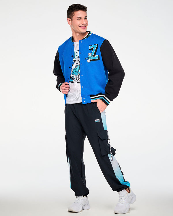 Male model with a hand in the pocket of the black cargo pants, showing the mesh side panel, the hip patch, and a 'ZUMBA' strap on the cargo pocket. Paired with a blue and black 'ZUMBA x THE SMURFS' varsity jacket.
