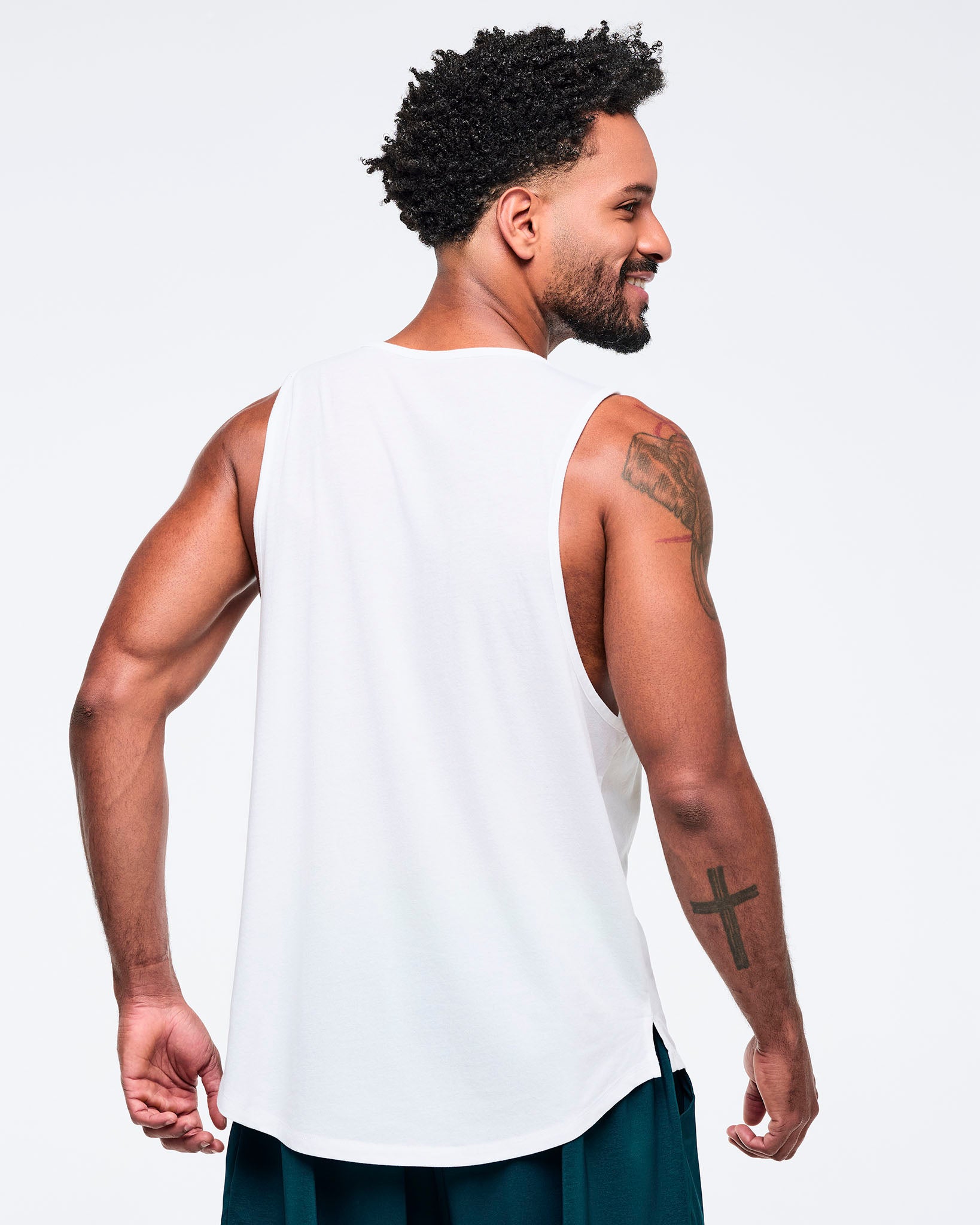Back view of the white racerback tank on a male model, showing the plain back and deep armholes, paired with teal pants.