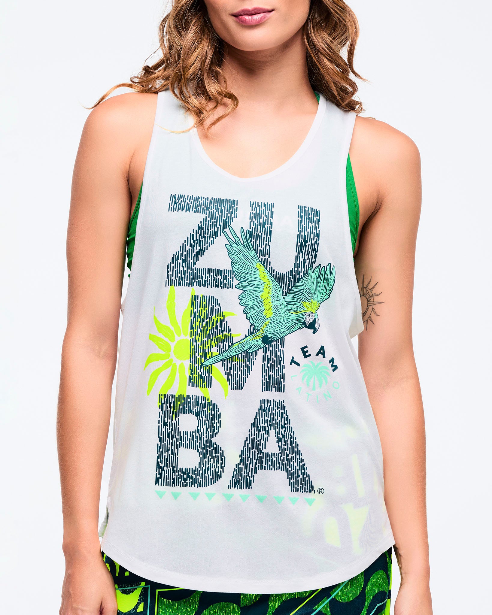 Close-up of the white racerback tank showing the “ZUMBA” text graphic with teal parrot and neon sunburst; a green sports bra is visible underneath on a female model.
