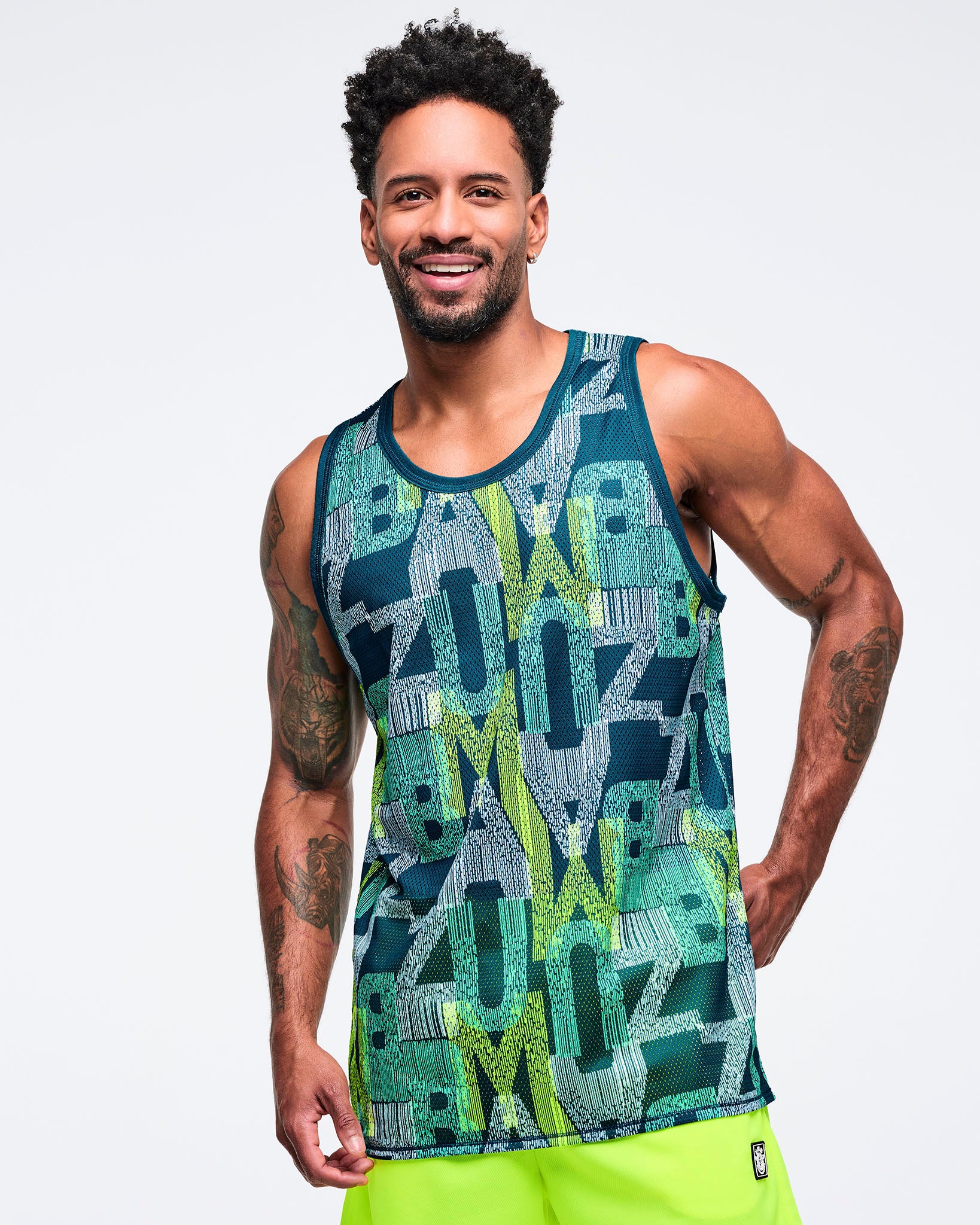 Teal, navy, and lime mesh tank top with allover “Zumba” graphic print and contrast binding at the crew neck and armholes, worn by a male model with neon lime shorts on a white studio background.