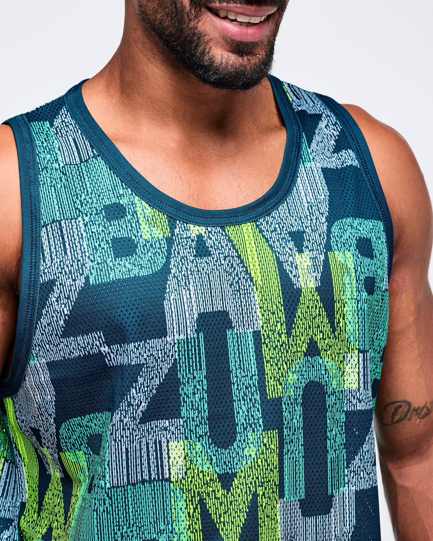 Close-up of the teal, navy, and lime “Zumba” graphic-print mesh tank top showing breathable mesh texture and contrast crew neckline on a male model.