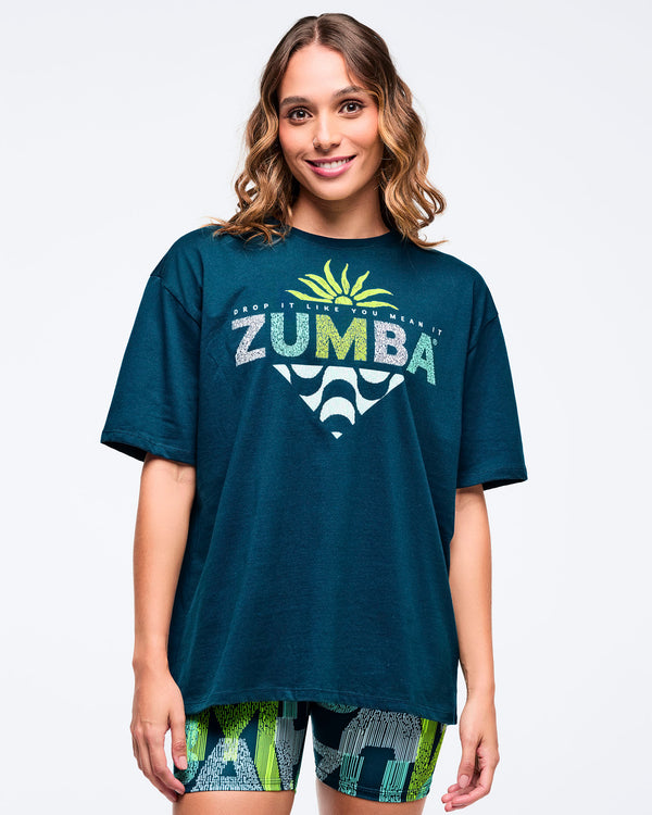 Dark teal oversized “ZUMBA” graphic T-shirt with lime sunburst and wave print, worn by a female model with green-and-blue patterned biker shorts.