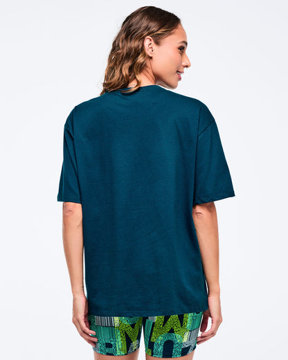 Back view of the dark teal oversized T-shirt on a female model, showing the clean solid back and loose sleeves, paired with green-and-blue patterned biker shorts.