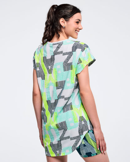 Back view of the longline all-over “ZUMBA” print cap-sleeve top on a female model, showing the curved hem and continuous pattern, paired with green-and-blue patterned biker shorts.