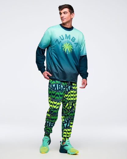Ombre aqua-to-deep teal graphic T-shirt with dark crewneck and palm tree “ZUMBA” chest print, layered over a dark long-sleeve top, worn by a male model with neon green-and-teal patterned joggers and aqua sneakers.