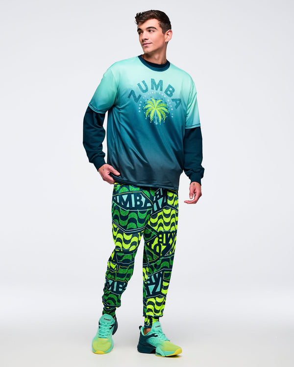 Ombre aqua-to-deep teal graphic T-shirt with dark crewneck and palm tree “ZUMBA” chest print, layered over a dark long-sleeve top, worn by a male model with neon green-and-teal patterned joggers and aqua sneakers.