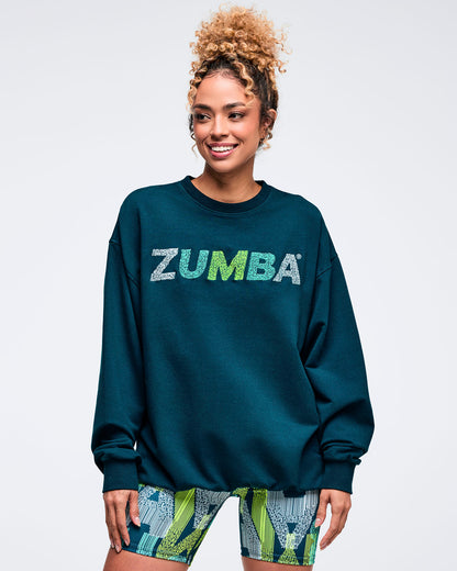 Teal crewneck sweatshirt with distressed “ZUMBA” chest logo, modeled by a female wearing teal-and-lime printed biker shorts on a white studio background.