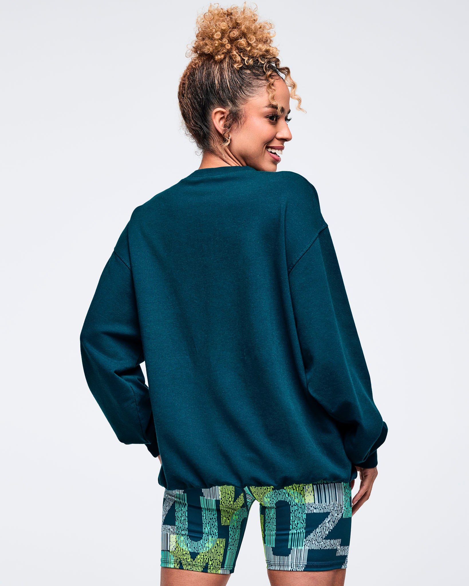 Back view of the teal crewneck sweatshirt with a loose, dropped-shoulder fit, modeled by a female wearing printed biker shorts on a white studio background.