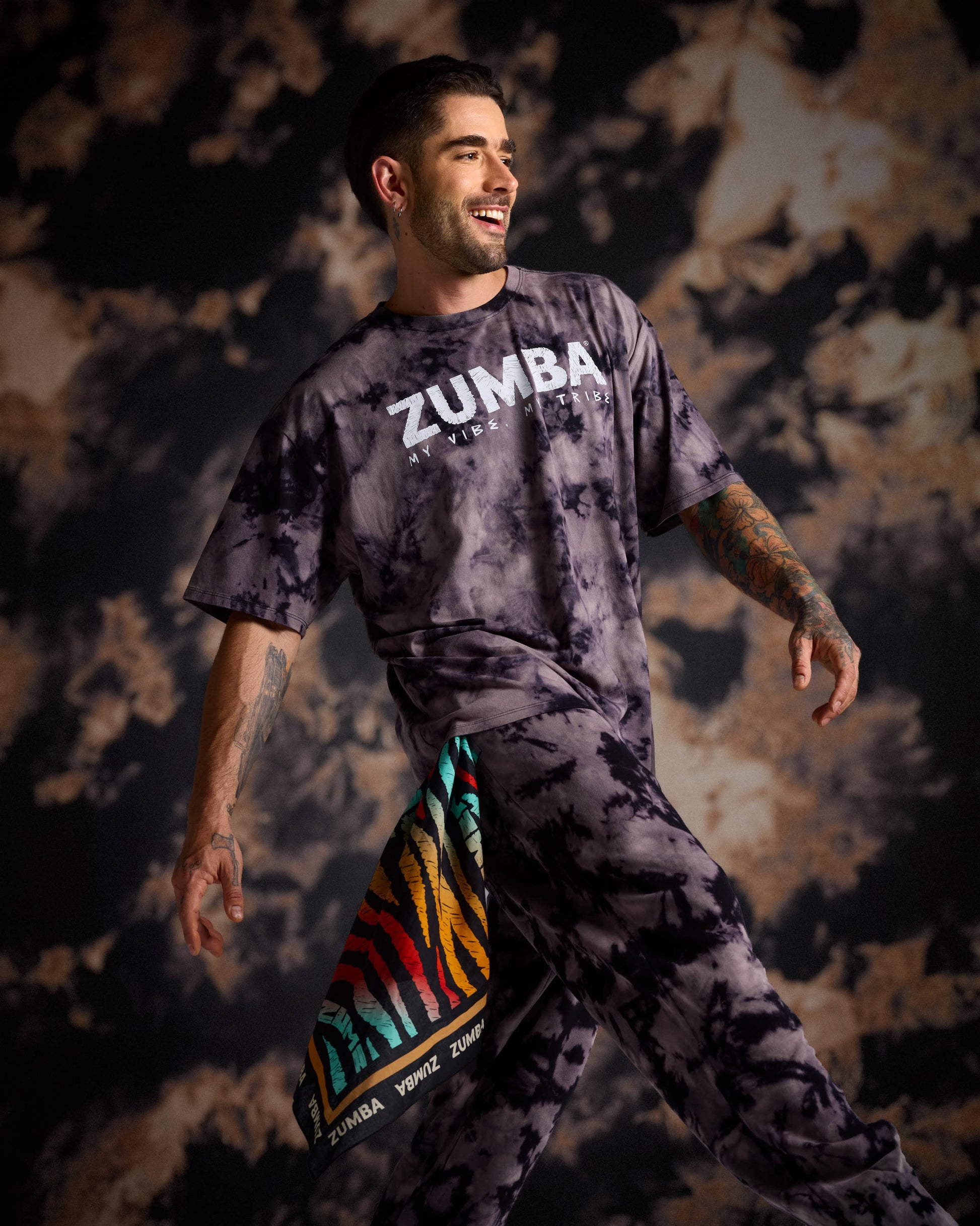 Male model in a black and tan tie-dye Zumba tee and matching pants with a multicolor bandana against a dark cloudy background.