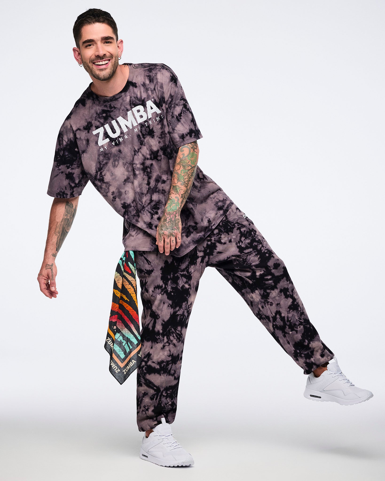 Oversized black and tan tie-dye Zumba T-shirt with a white “ZUMBA My Vibe. My Tribe.” graphic on the front, paired with matching tie-dye sweatpants. Male model wears the loose-fit short-sleeve crewneck tee, coordinating sweatpants, a colorful side bandana with white sneakers.