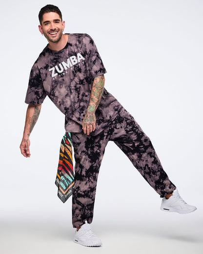 Oversized black and tan tie-dye Zumba T-shirt with a white “ZUMBA My Vibe. My Tribe.” graphic on the front, paired with matching tie-dye sweatpants. Male model wears the loose-fit short-sleeve crewneck tee, coordinating sweatpants, a colorful side bandana with white sneakers.