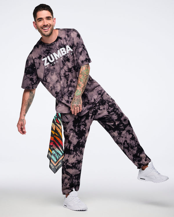 Oversized black and tan tie-dye Zumba T-shirt with a white “ZUMBA My Vibe. My Tribe.” graphic on the front, paired with matching tie-dye sweatpants. Male model wears the loose-fit short-sleeve crewneck tee, coordinating sweatpants, a colorful side bandana with white sneakers.