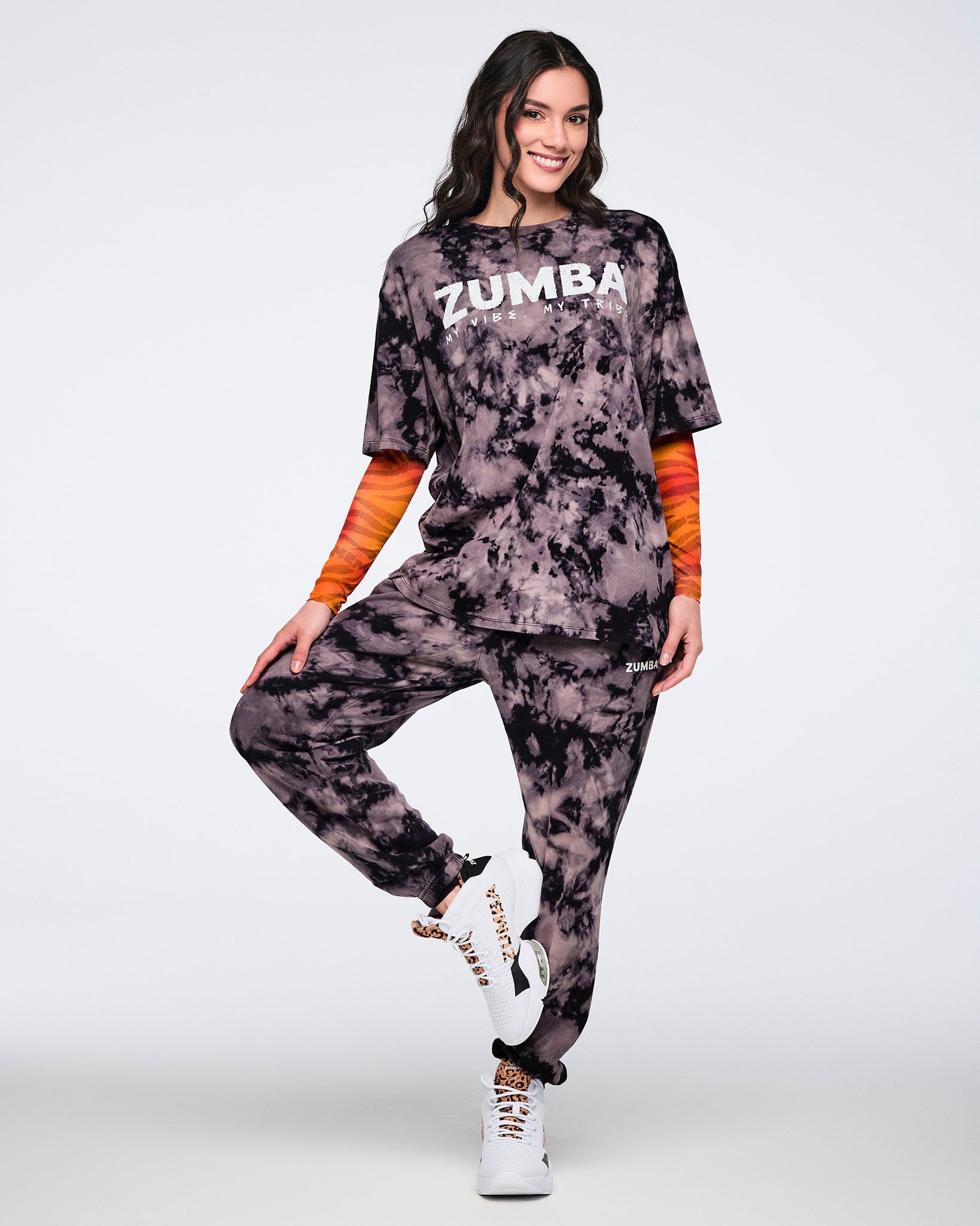 Oversized black and tan tie-dye Zumba T-shirt with a white front graphic, paired with matching tie-dye sweatpants. Female model wears the short-sleeve crewneck tee over a fitted orange long-sleeve top, styled with white sneakers.