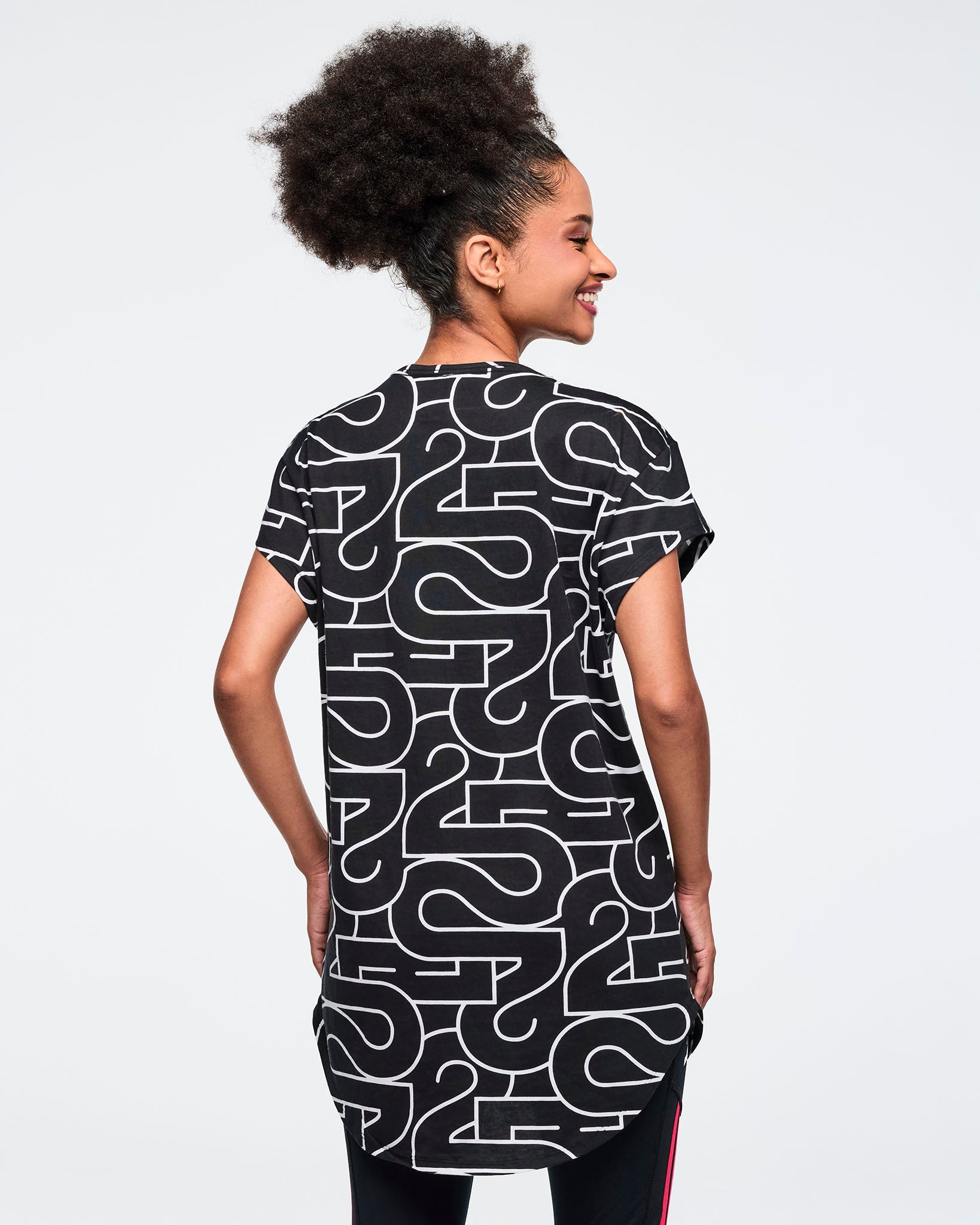 Black, long, round hem tee with an allover white 25 graphic, shown from the back. A smiling female model wears the short-sleeve, round hem tee over black leggings against a light gray background.