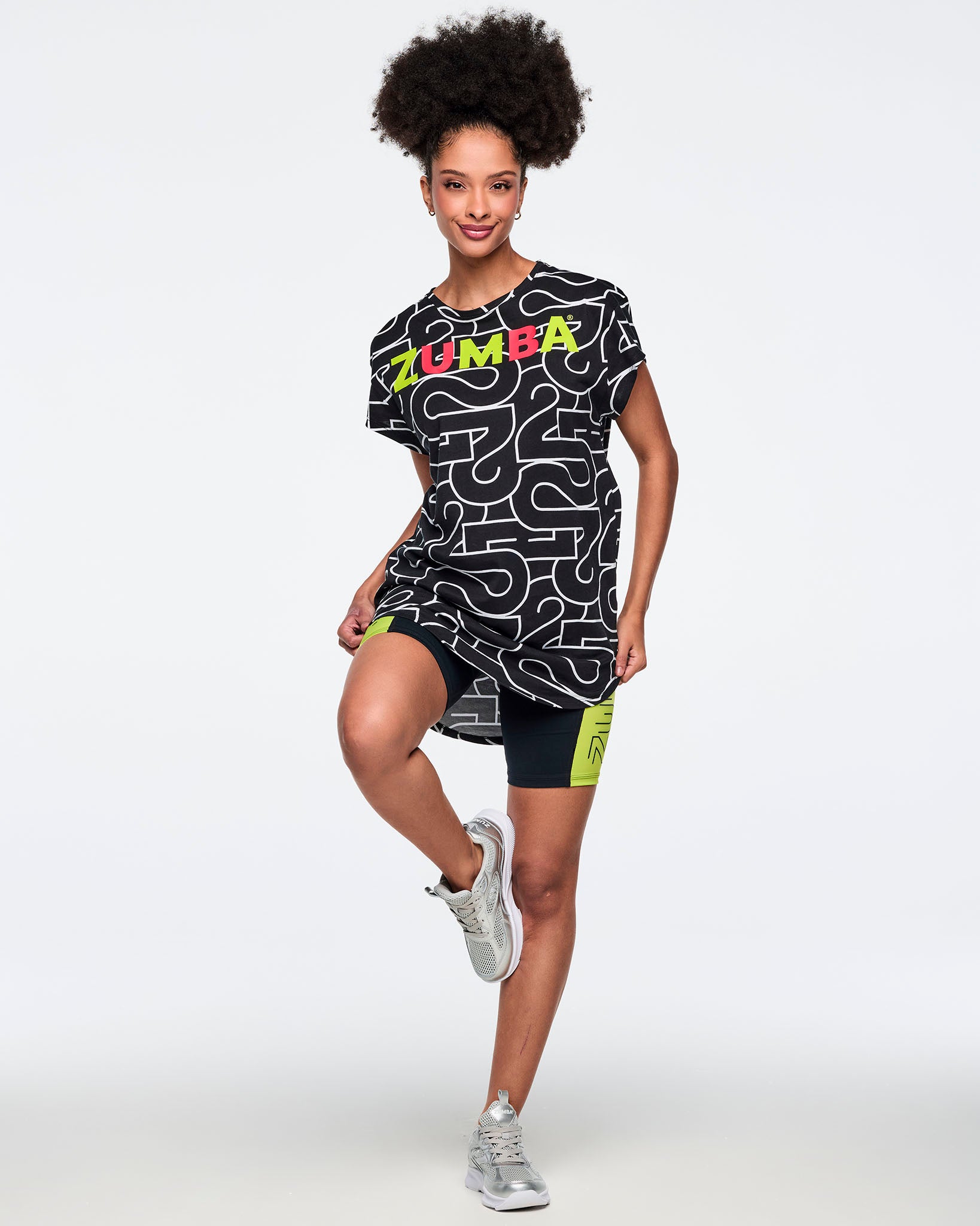 Black, long, round hem tee with an allover white 25 graphic and multicolor Zumba logo across the chest. A female model wears the round hem tee, black bike shorts with neon green side panels and silver sneakers against a light gray background.