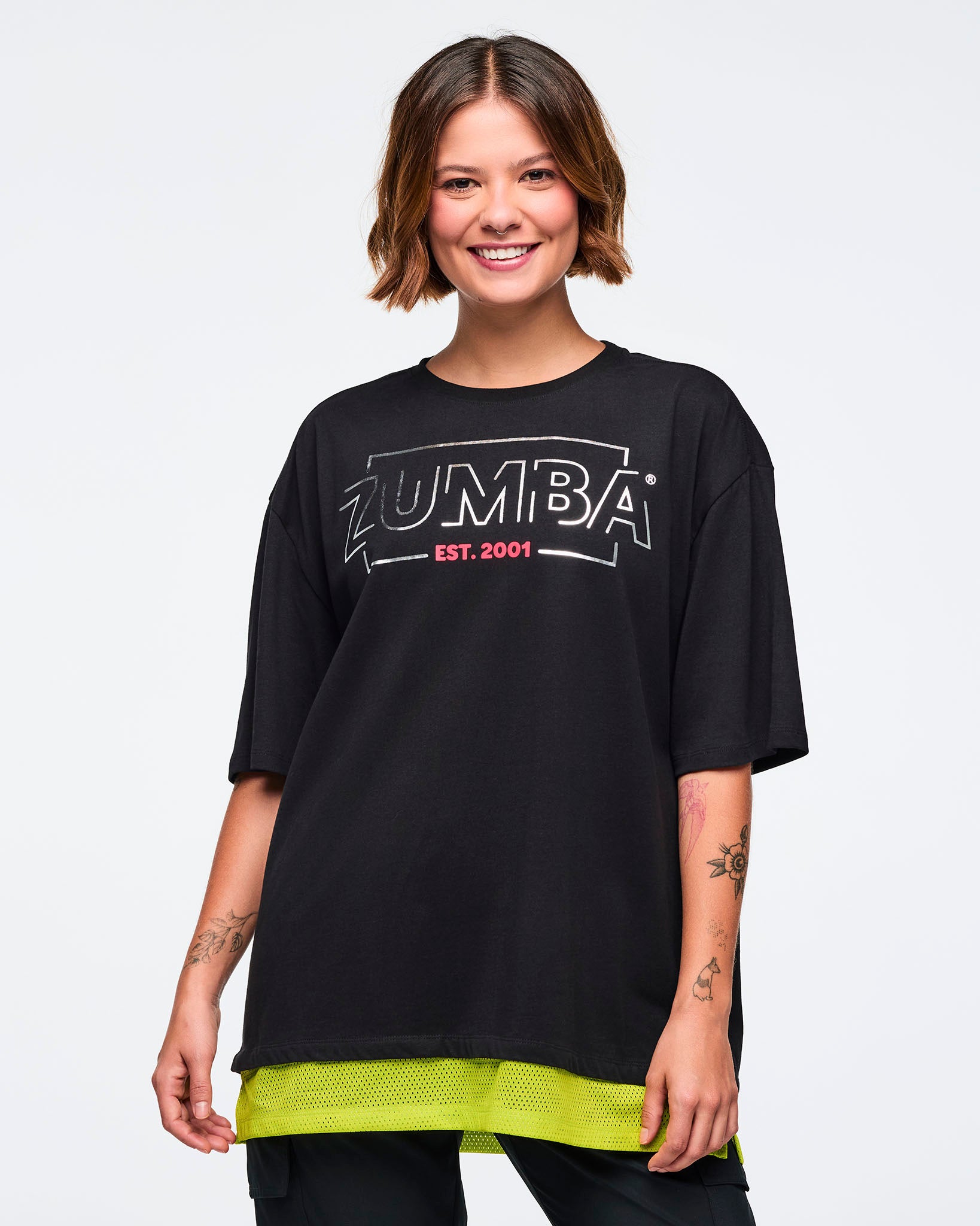 Black oversized Zumba T-shirt with a large silver ZUMBA logo and pink “EST. 2001” text on the front, with an extended double layered mesh hem panel. Female model is smiling and wearing the loose short-sleeve tee with black bottoms.