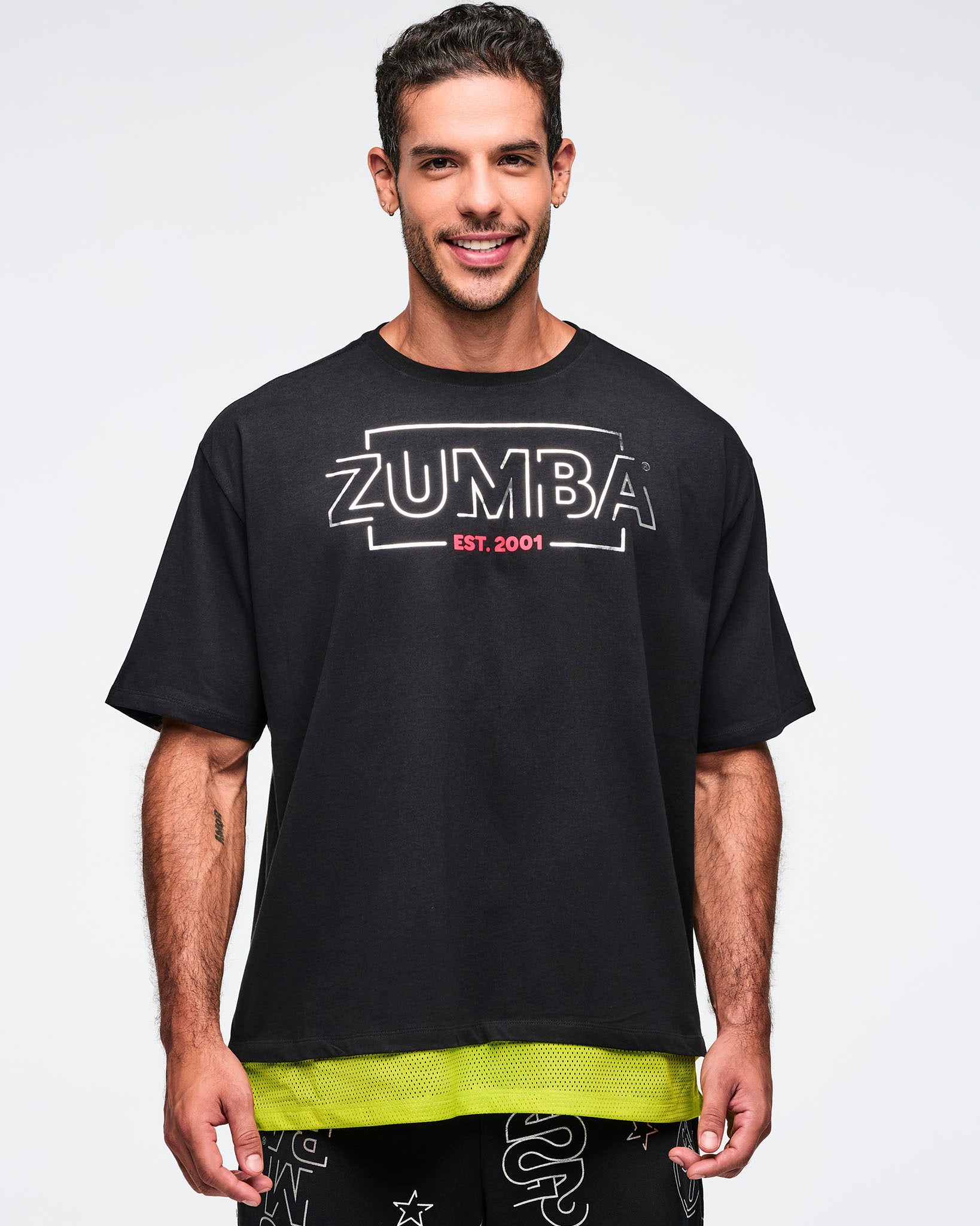 Black oversized Zumba T-shirt with a large silver ZUMBA logo and pink “EST. 2001” text on the front, with an extended double layered mesh hem panel. Male model is wearing the loose short-sleeve tee with black graphic pants.