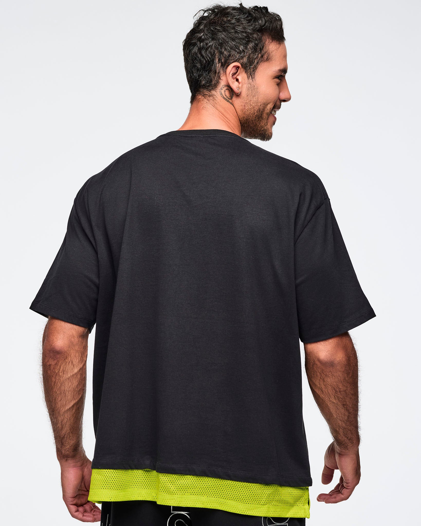 Black oversized Zumba T-shirt shown from the back, with an extended double layered mesh hem panel. Male model is turned away and wearing the loose short-sleeve tee with black graphic pants.