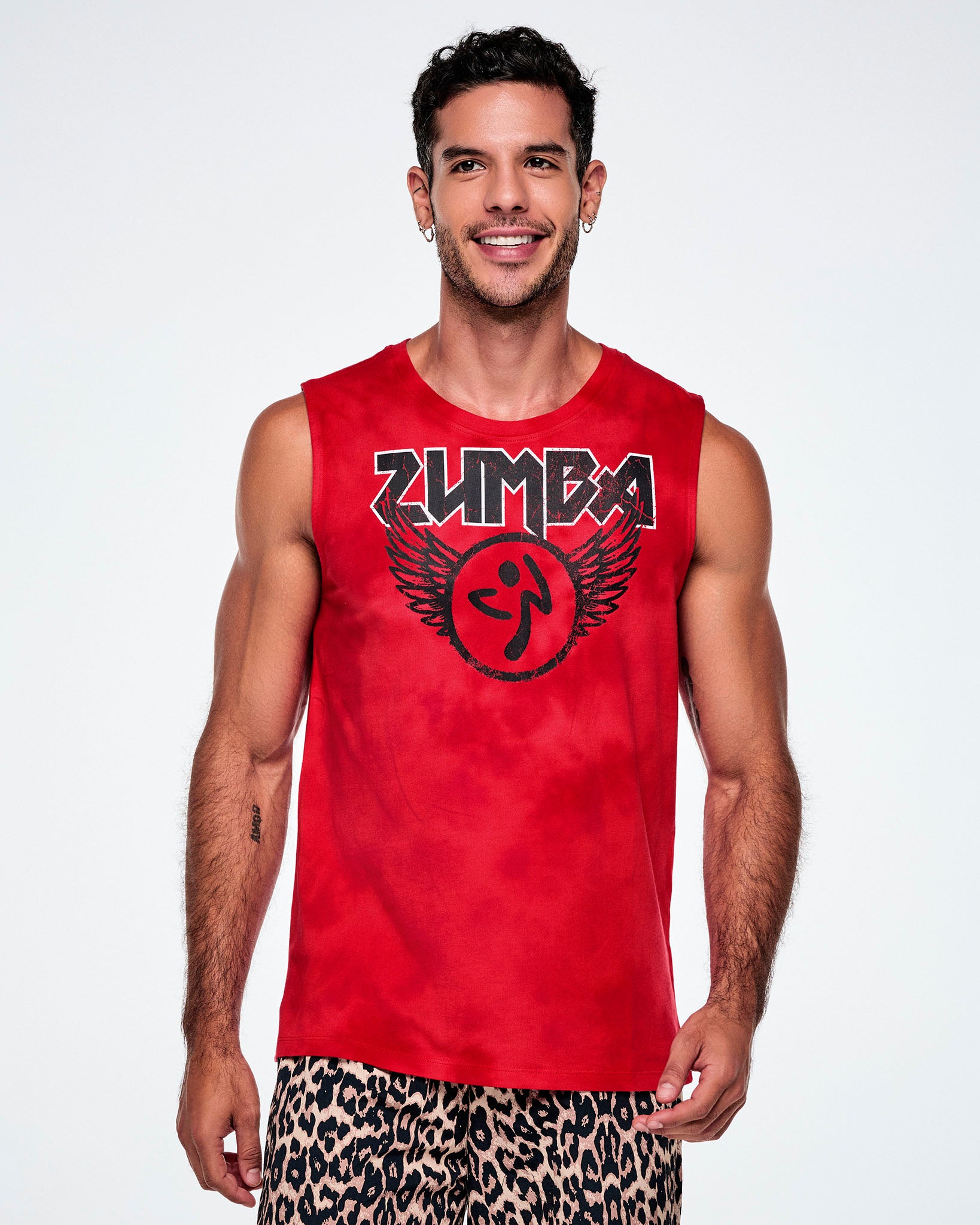 A male model wearing a red tank top with a distressed black and white Zumba text with a large black Zumbito graphic with wings, paired with tan leopard print shorts.