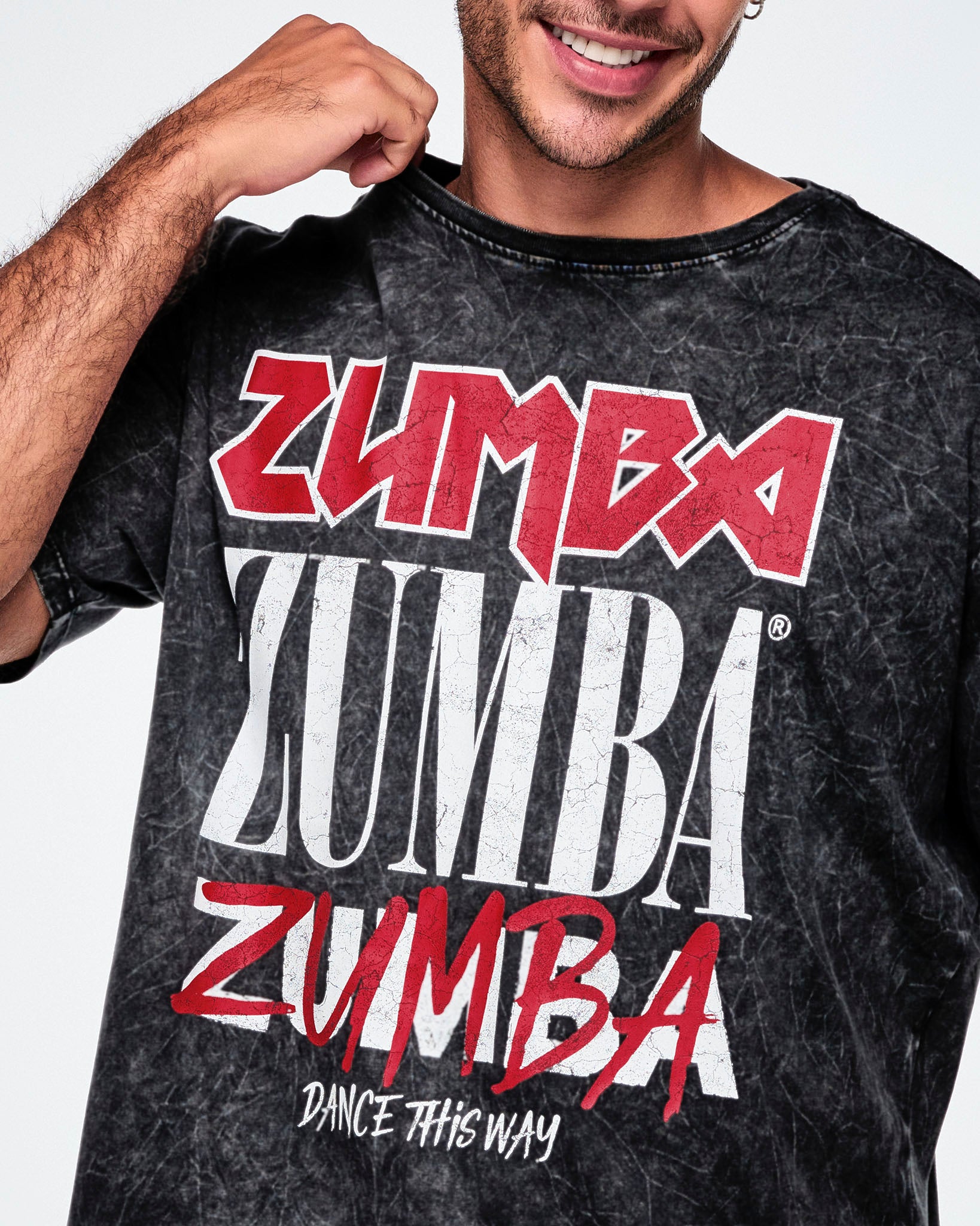 A close-up of a male model wearing a black acid-wash graphic tee featuring the word 'ZUMBA' printed in red and white multiple times, with 'DANCE THIS WAY' underneath.