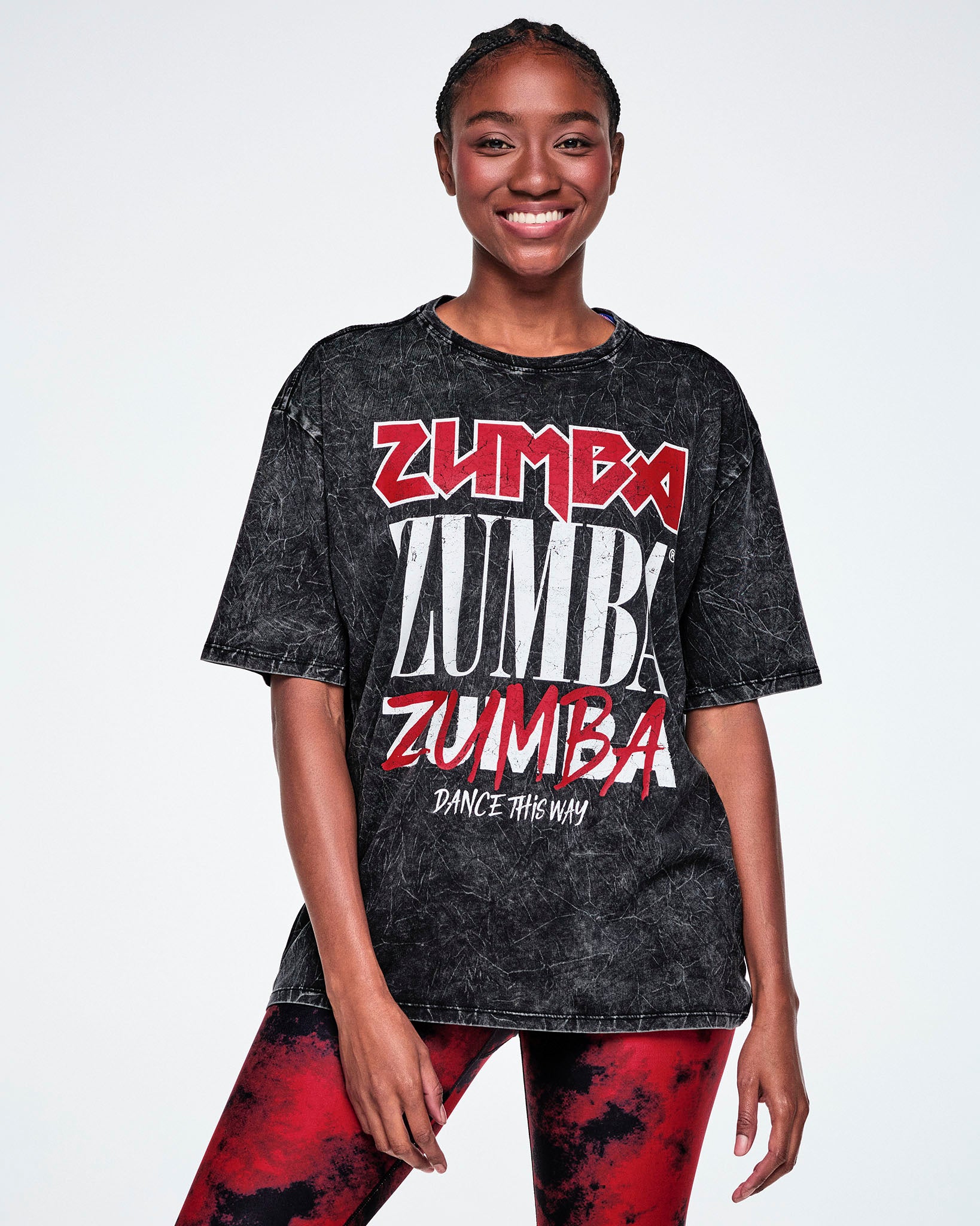 A female model wearing a black acid-wash graphic t-shirt with 'ZUMBA' printed in red and white in four different fonts, with "Dance This Way" in white text below and paired with red and black leggings.