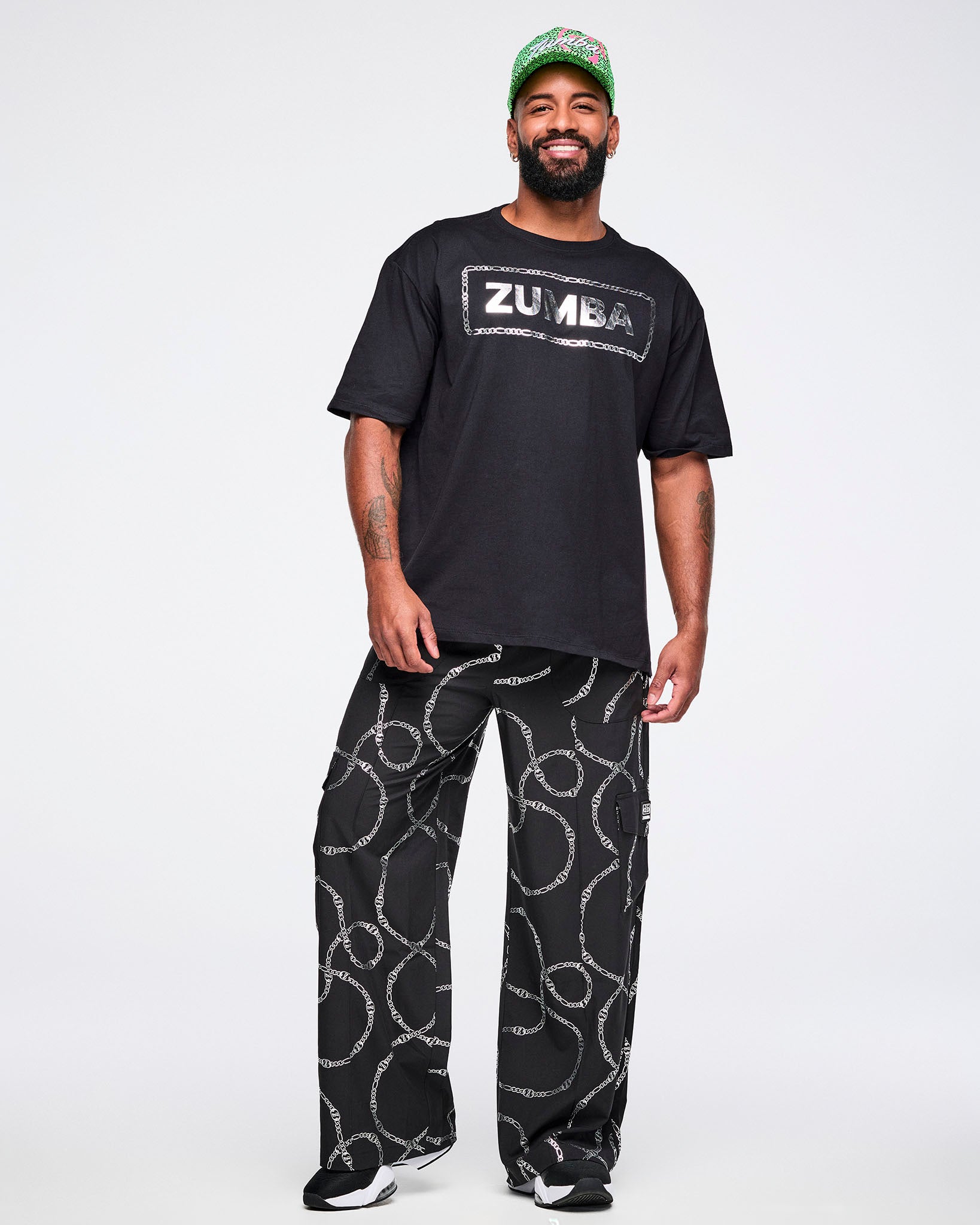 Full view of a male model wearing a black Zumba T-shirt with “ZUMBA” in a silver patterned rectangle, paired with matching pants and black and white shoes.