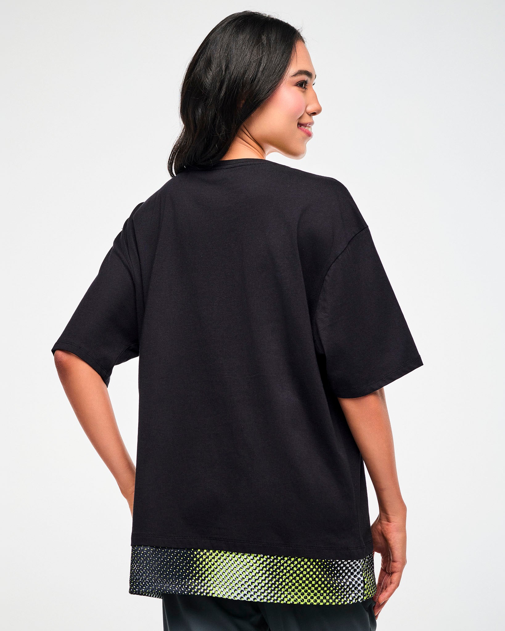 Back view of a female model wearing a black oversized T-shirt with a yellow, black, and white mesh dot-print hem.