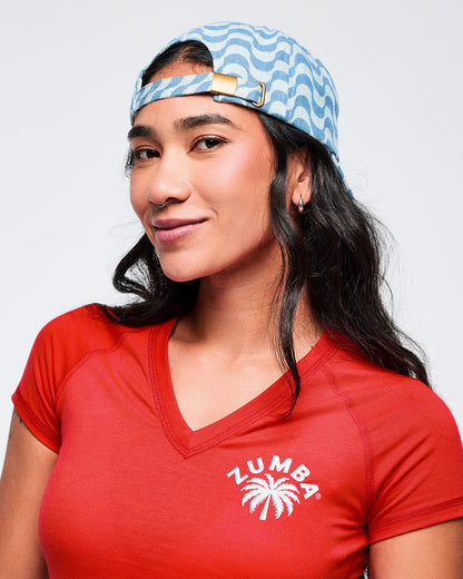 Light blue wavy-print baseball cap worn backward, showing the adjustable strap and metal buckle, modeled by a female wearing a red V-neck tee on a white studio background.