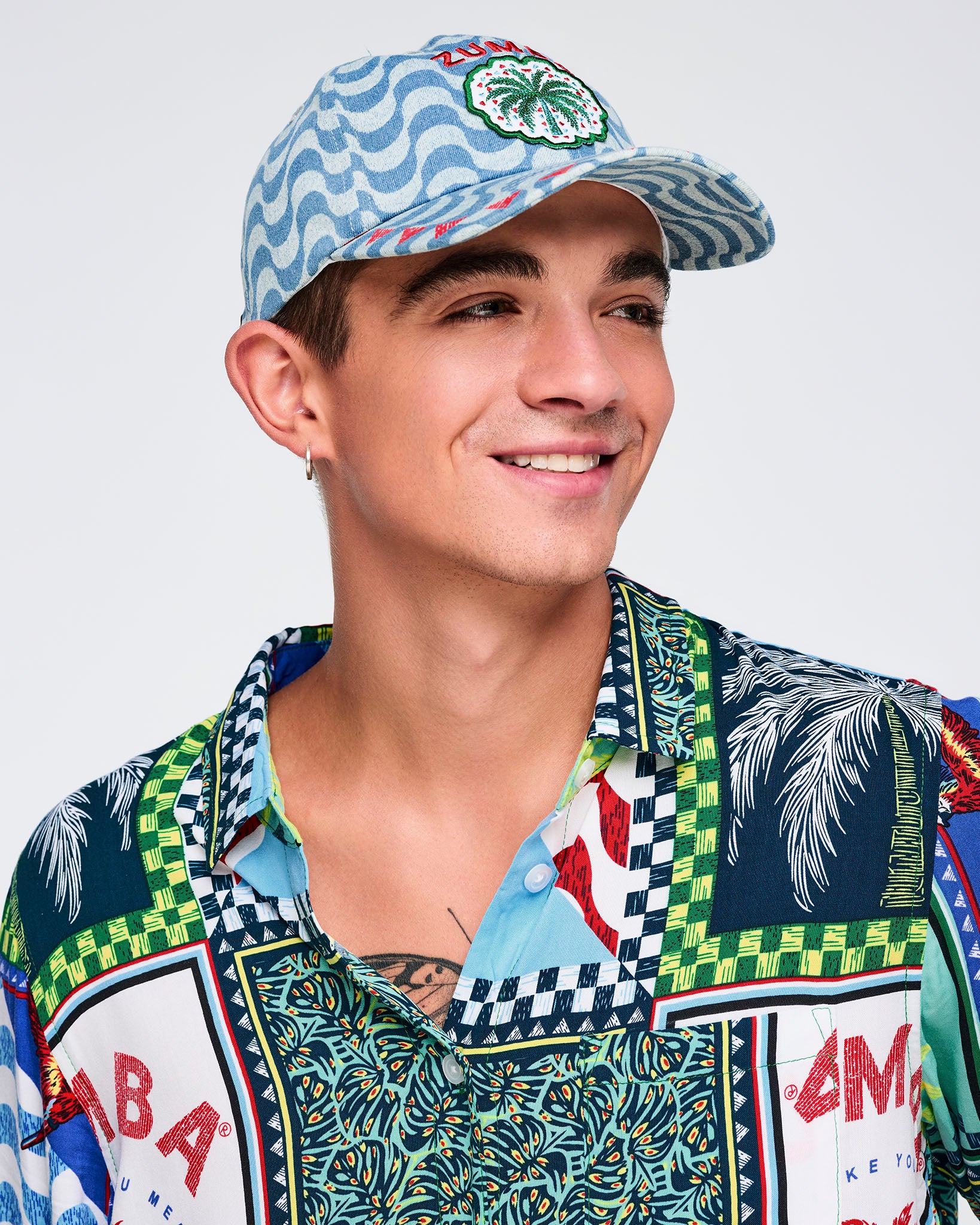 Light blue wavy-print baseball cap with embroidered “ZUMBA” text and palm-tree patch, modeled by a male wearing a multicolor printed button-up shirt on a white studio background.