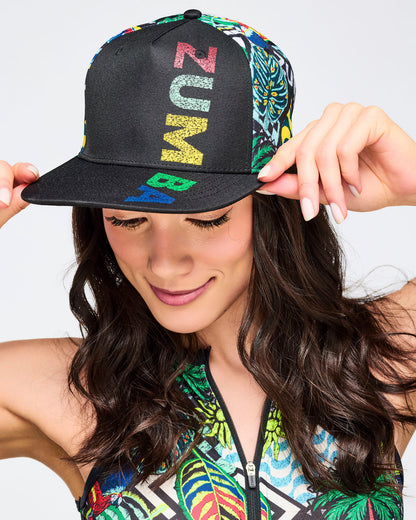 Close-up of a woman wearing a black snapback cap with “ZUMBA” printed vertically in multicolor letters on the front panel; the side panels have a bright tropical/graphic print. She’s holding the brim with both hands.