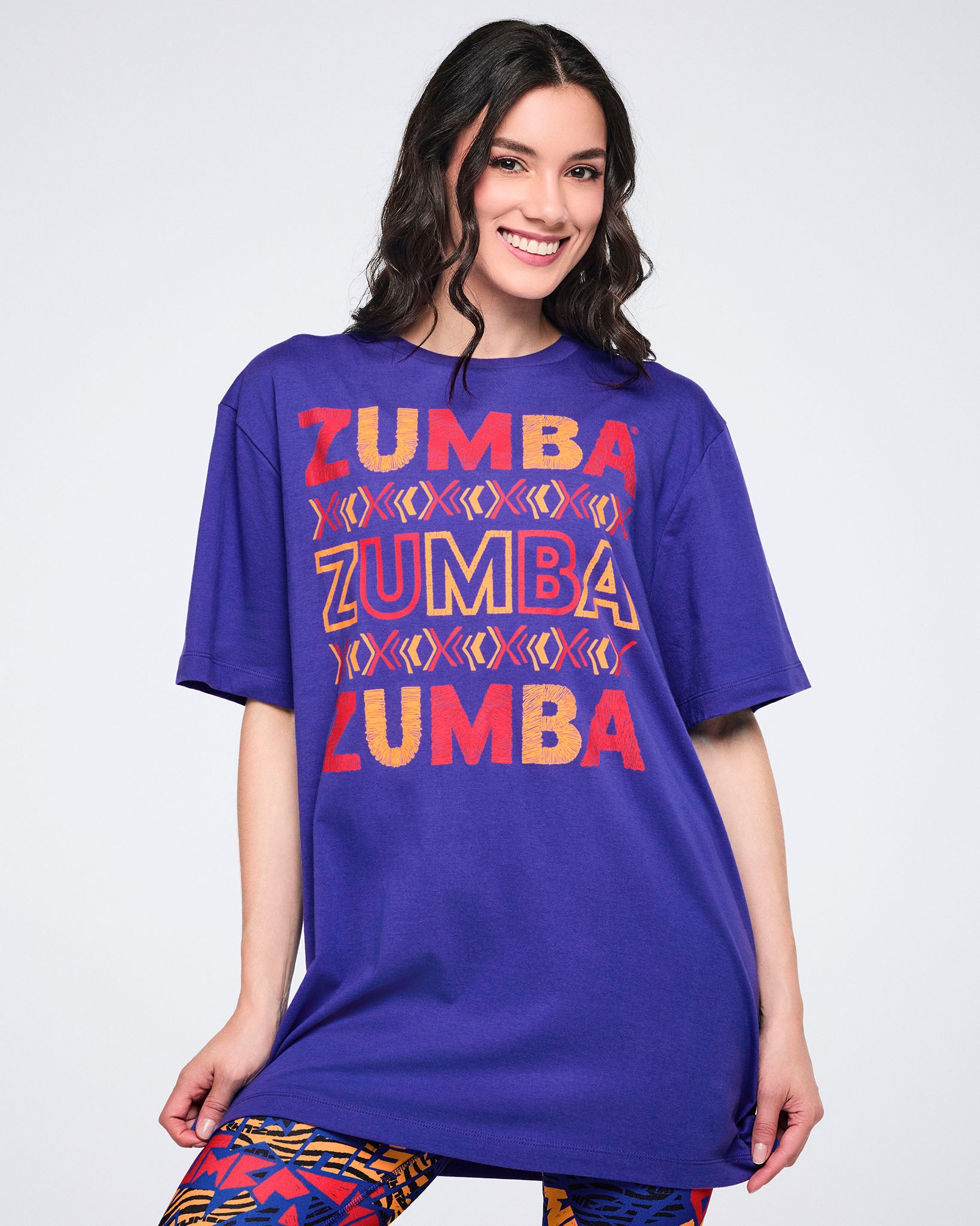 Female model wearing an oversized purple short-sleeve Zumba T-shirt with stacked red and orange logo graphics, paired with blue, orange, and black patterned leggings.
