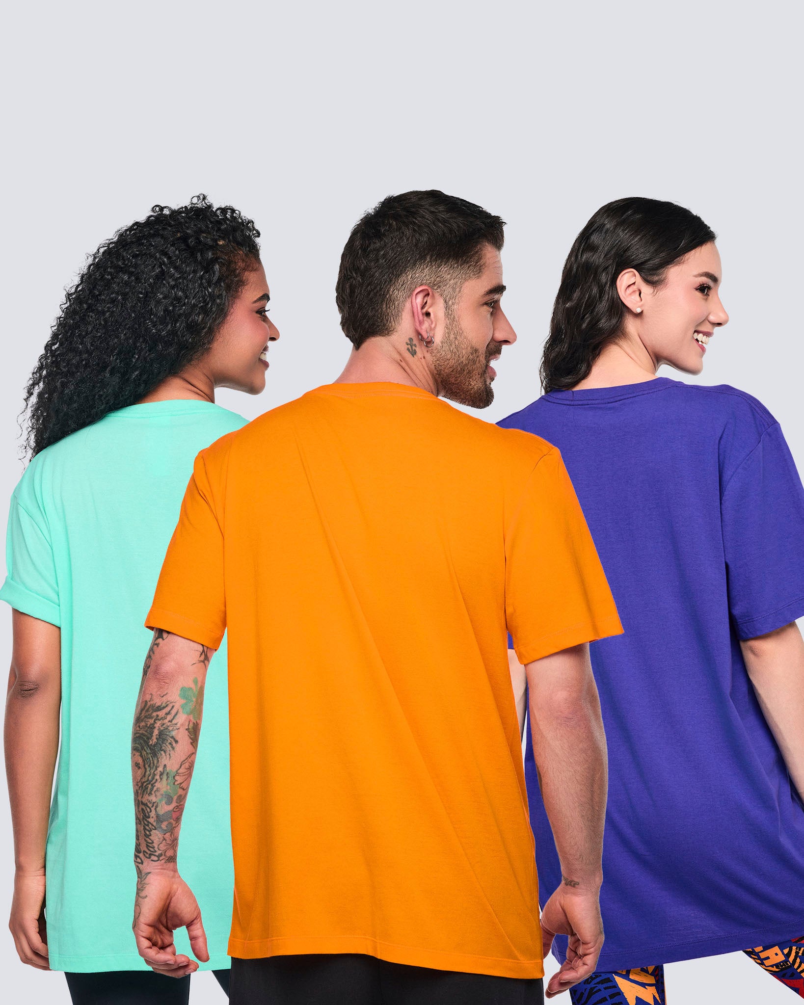 Male and female models shown from the back wearing oversized graphic Zumba T-shirts in orange, mint, and purple with short sleeves and relaxed fits, styled with black leggings or printed bottoms.