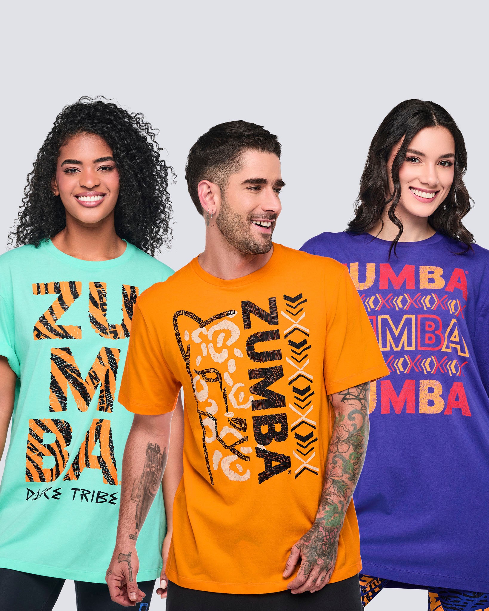 Male model wearing an orange crewneck T-shirt with a bold black vertical Zumba graphic and abstract animal-print design, alongside female models in oversized mint and purple graphic Zumba T-shirts, shown from the front.