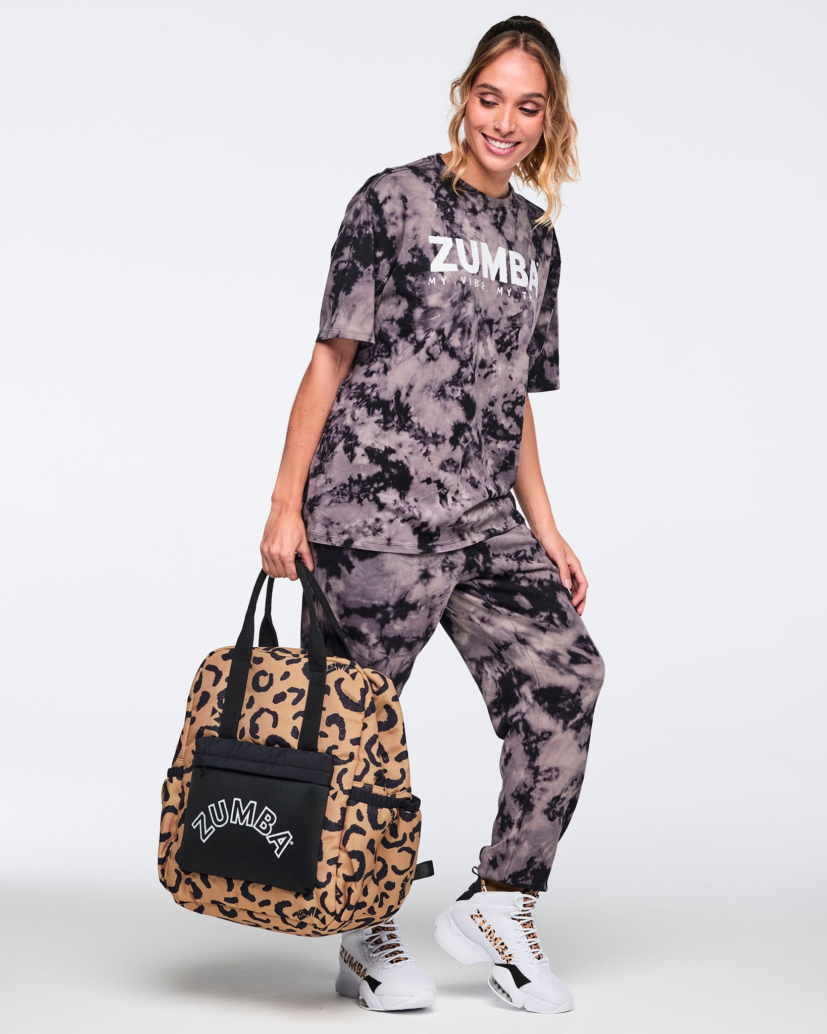 Leopard-print Zumba backpack with black handles and a black front pocket with white ZUMBA logo, carried by hand. Female model wears a loose black and gray tie-dye Zumba T-shirt, matching tie-dye joggers, and white Zumba sneakers with leopard accents while posing against a light gray background.