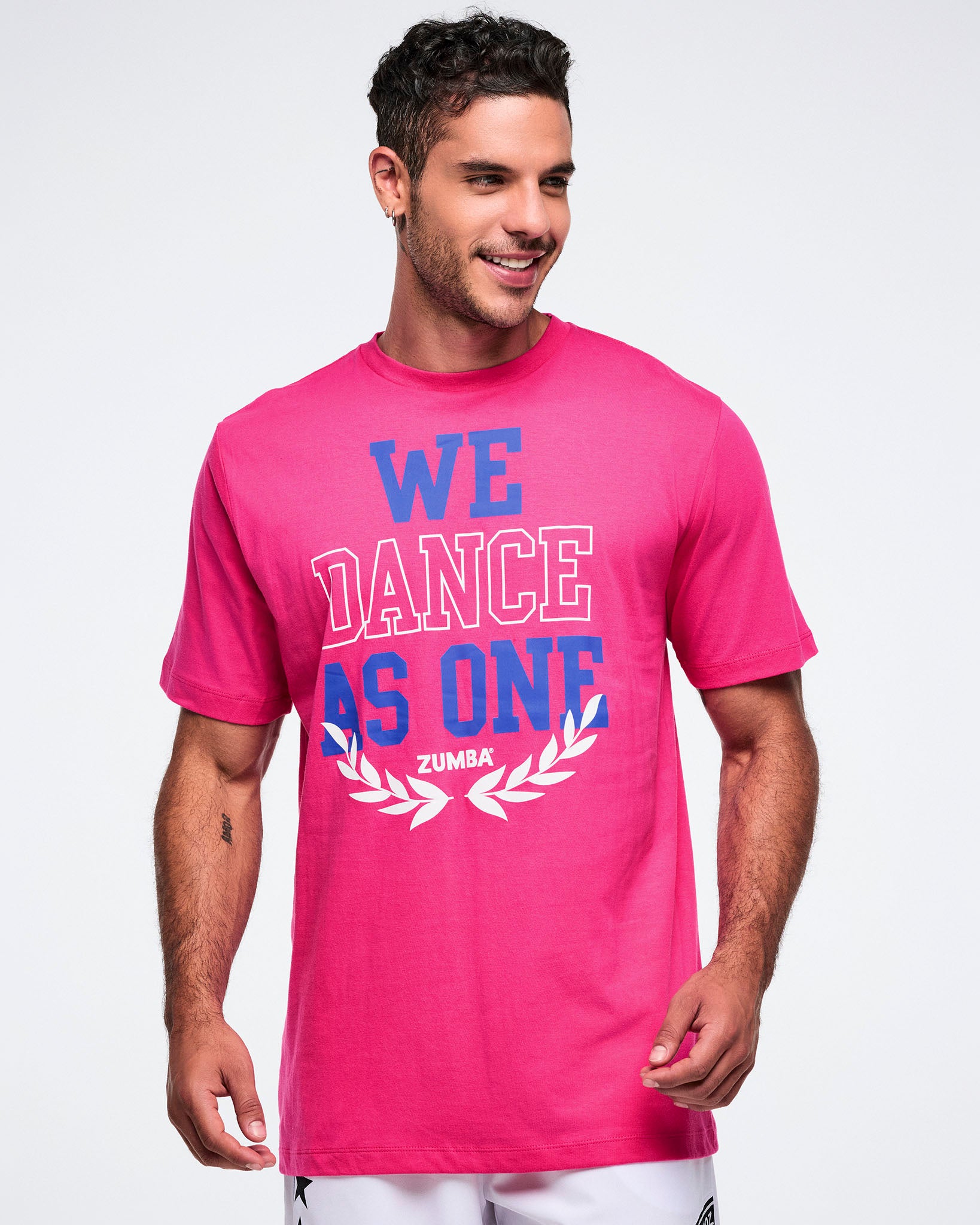 Male model wearing a pink Zumba t-shirt with 'We Dance As One' text on a white background.