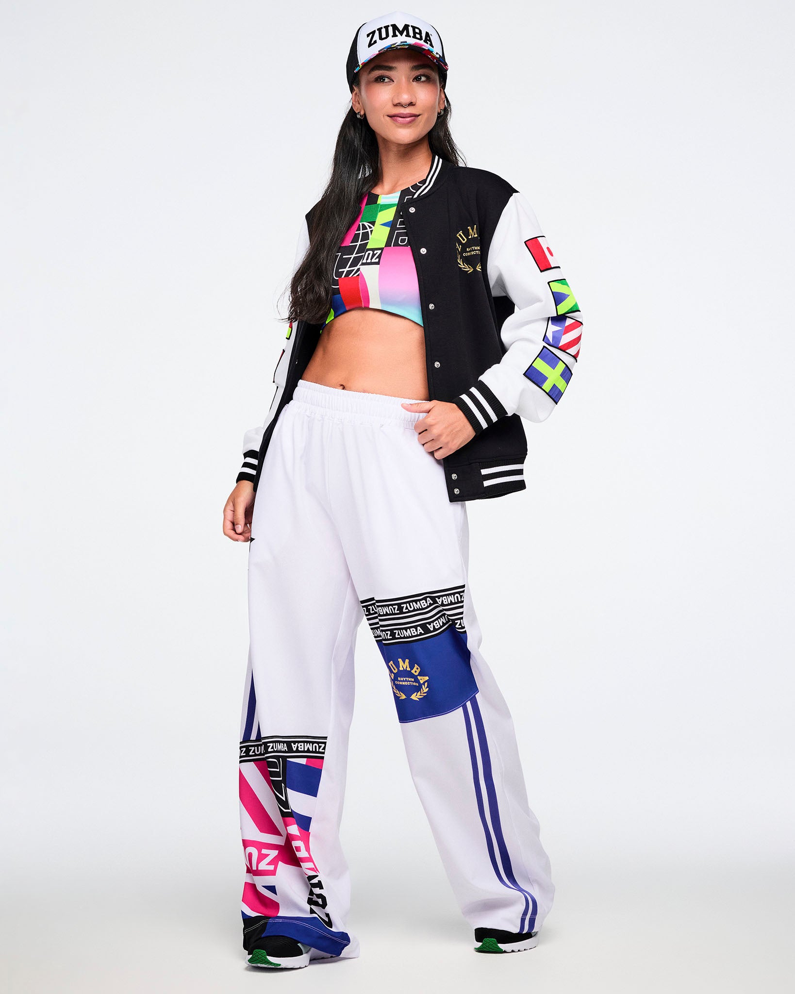 Female model wearing a hat, colorful bra, a black and white varsity jacket with white pants on a white background.