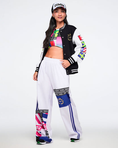 Female model wearing a hat, colorful bra, a black and white varsity jacket with white pants on a white background.