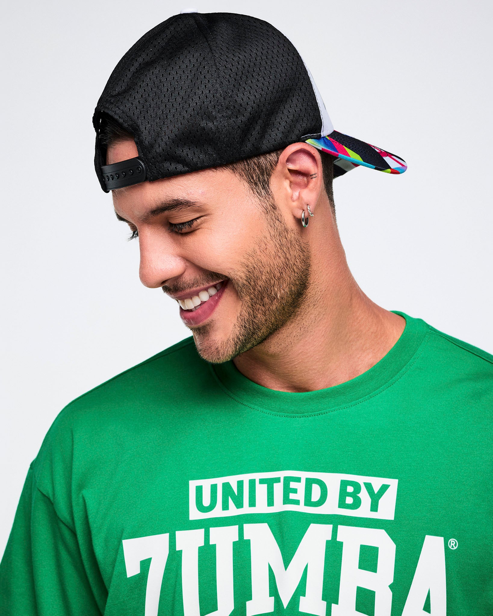 Male model wearing a green 'United by Zumba' shirt and a backwards black cap with a colorful bill.