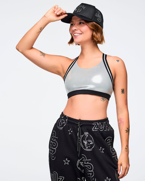 Black baseball cap with white Zumba logo on the front and a silver 25 Years graphic on the side. A smiling female model wears the cap with a shiny light gray sports bra with black trim and loose black joggers printed with silver Zumba graphics against a light gray background.