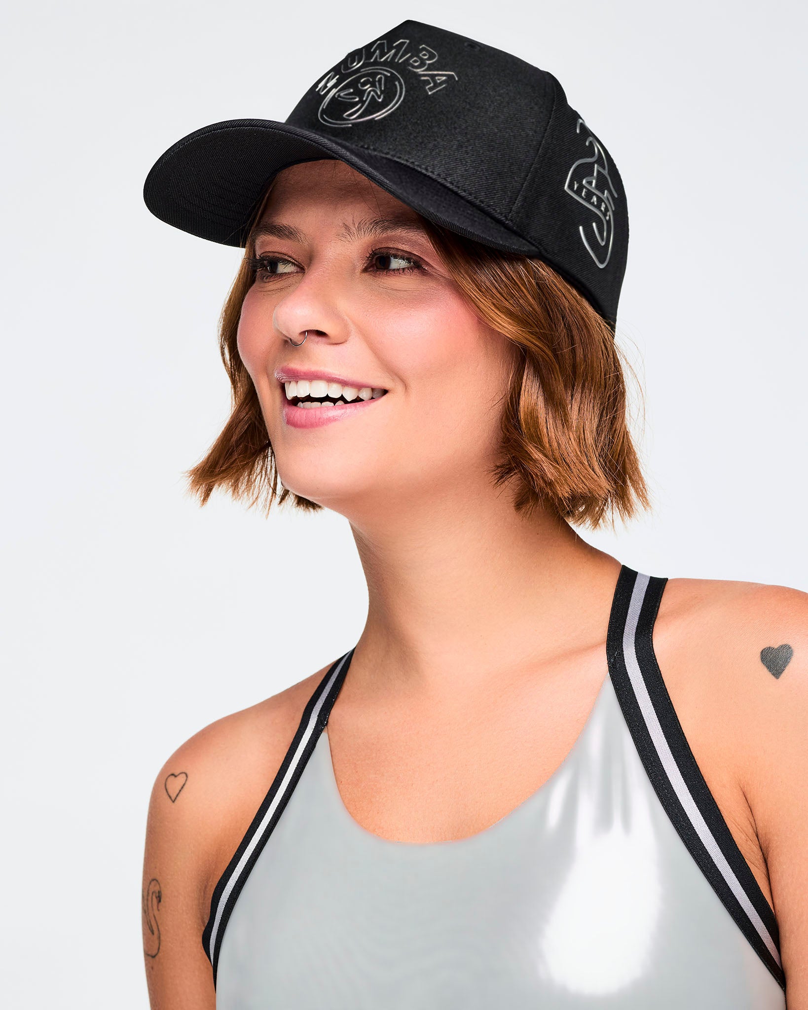 Black baseball cap with silver Zumba logo on the front and a white 25 Years graphic on the side, shown in a close-up lifestyle image. A smiling female model wears the cap with a shiny silver sports bra with black trim against a light gray background.