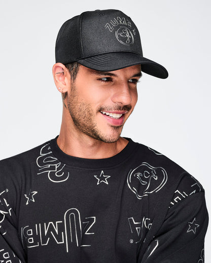 Black baseball cap with a curved brim and tonal Zumba logo on the front. A smiling male model wears the cap with a black crewneck sweatshirt covered in silver Zumba graphics against a light gray background.