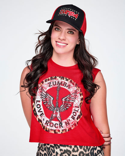 A female model wearing a black hat with red mesh side panels and a red embroidered ZUMBA logo outlined in silver studs on the front and paired with a red sleeveless crop top with a large distressed Zumba logo graphic on the front and tan leopard print leggings.