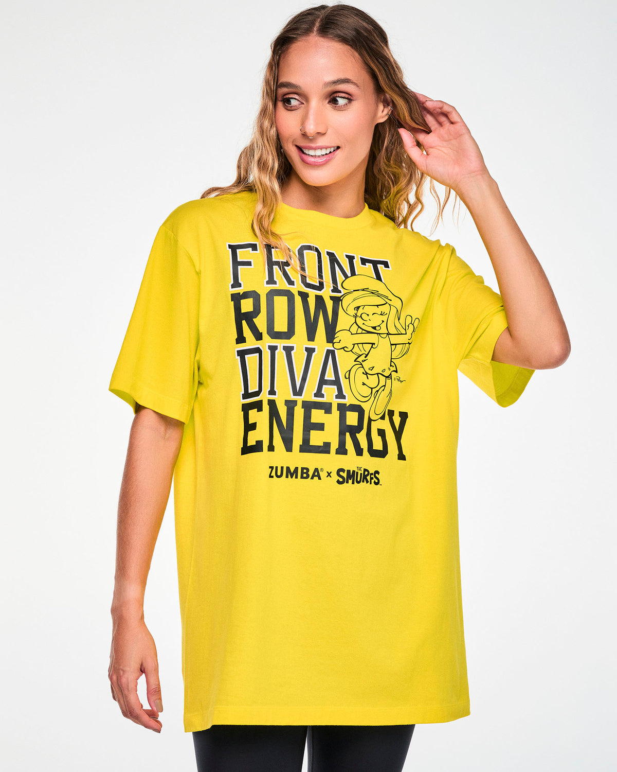 Female model in a yellow crew neck tee with stacked black varsity 'FRONT ROW DIVA ENERGY' text, a black line drawing of a winking Smurfette beside the words, and 'ZUMBA' X 'THE SMURFS' logos below. Paired with black leggings.