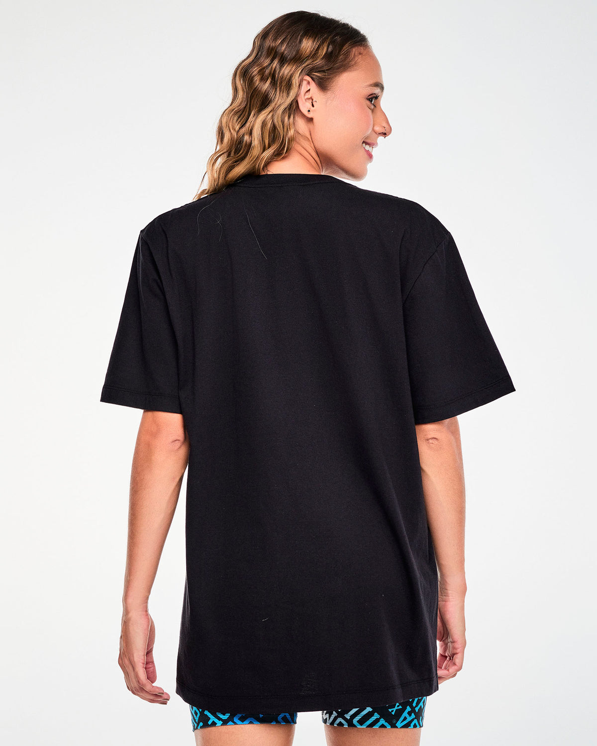 Back view of a female model in the black 'ZUMBA' and 'THE SMURFS' tee, showing a plain back and an easy, long fit.