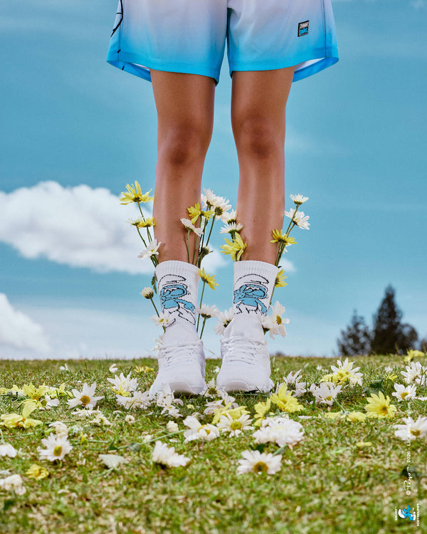 Close-up of a female model's legs in the Smurf crew socks and white shoes, with daisies tucked into the socks and scattered on the grass, and navy-to-light-blue ombre mesh shorts at the top of the frame, all against a blue cloudy sky.