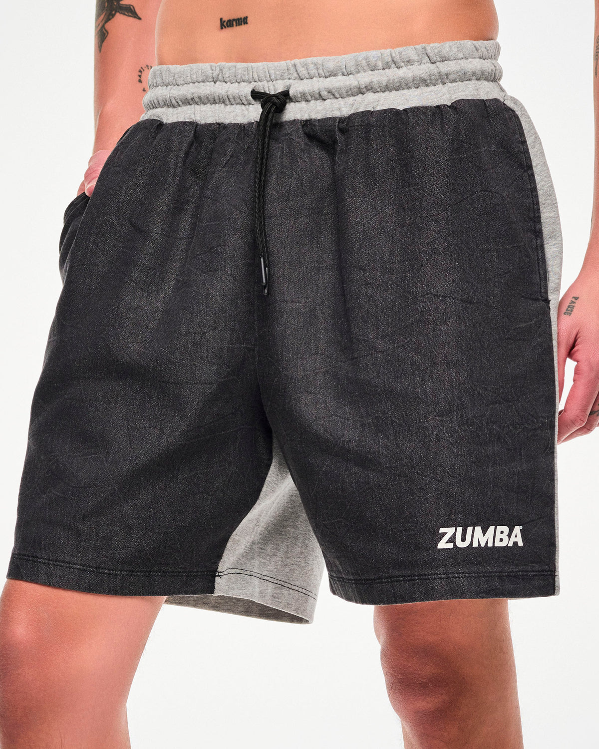 Male model wearing black denim and grey shorts with a drawstring waist, white "ZUMBA" text on the lower left hem, and white sneakers, hand in the side pocket.