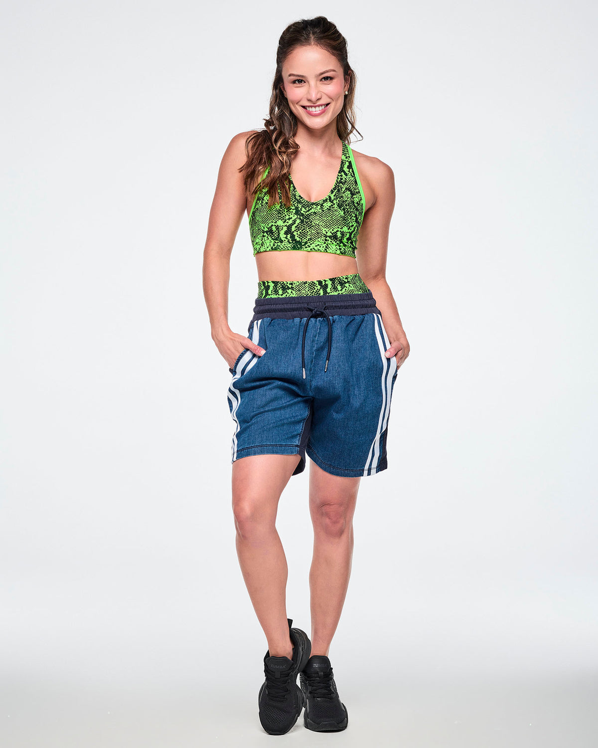 Female model wearing a neon green snake-print bra top with denim combo shorts featuring a navy back panel and double white stripe, paired with black sneakers.