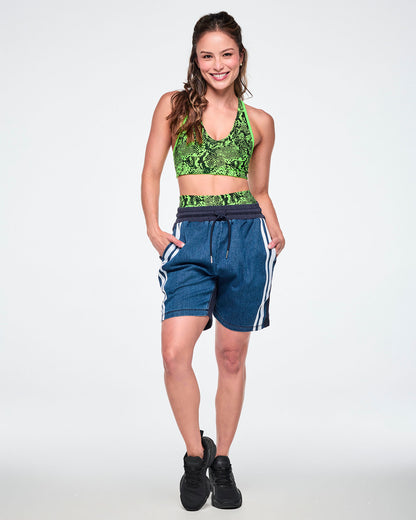 Female model wearing a neon green snake-print bra top with denim combo shorts featuring a navy back panel and double white stripe, paired with black sneakers.