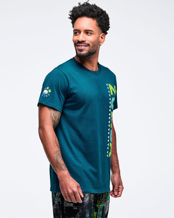 Teal crewneck T-shirt with short sleeves and a vertical lime “ZUMBA” graphic along the side front, shown in side view on a male model wearing patterned shorts on a white studio background.